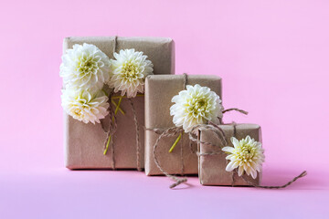 Three gift boxes in craft paper with white dahlia flowers on a gradient purple-pink background. The concept of a holiday, Valentine's day, women's day, birthday, wedding, birthday