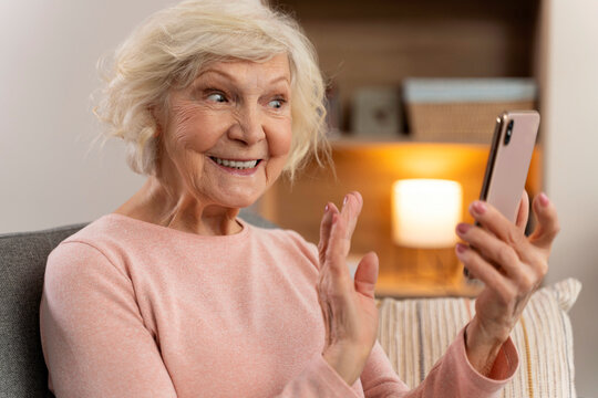 Happy mature old woman using mobile phone app for video call, waving hand, feeling excited while sitting on couch at home. Stock photo
