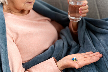 Cropped view of the senior woman holding glass of water and looking on pills while laying at the sofa at home. Age, medicine, healthcare and people concept