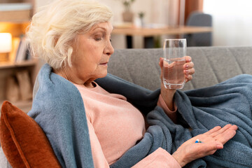 Senior woman holding glass of water and looking on pills while laying at the sofa at home. Age, medicine, healthcare and people concept