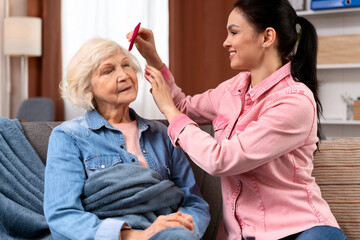 Brunette daughter helping senior woman to comb hair and make hairstyle when sitting on sofa at home together. Stock photo