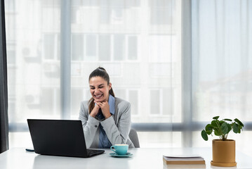 Young happy satisfied successful businesswoman manager and executive sitting in office casually working on laptop computer enjoying working day self-conscious and grateful for success and hard work