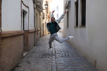 Obraz na płótnie Canvas A handsome young man with a beard and glasses is jumping for joy in a narrow street. Happiness and joy concept.