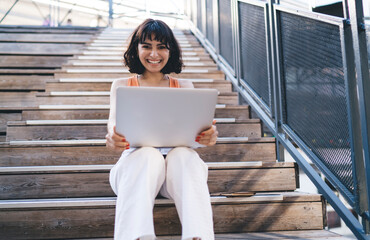 Smiling woman sitting on wooden stairs with laptop in hands