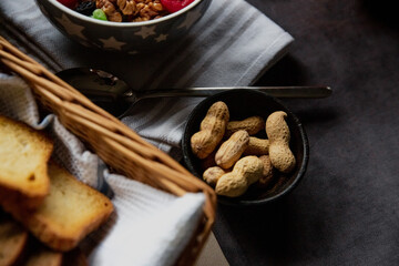 Rustic breakfast setting with a bowl of granola and berries, whole peanuts in a dish, toast in a basket, and a spoon on linen. A cozy flat lay with natural textures and warm morning light.
