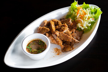 Delicious Thai-style grilled pork with chili powder and roasted rice served with Thai style dipping sauce and lettuce on a white plate on a black table.