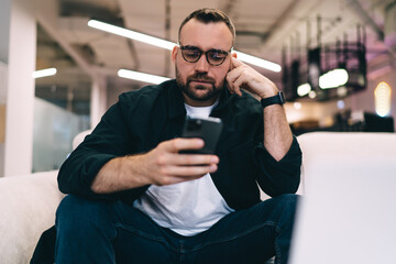 Man in glasses messaging on smartphone while sitting on armchair