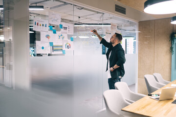 Busy male entrepreneur drawing scheme with marker on glass wall