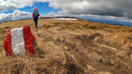 Woman walking next to path mark with Austrian flag painted on rock on remote alpine meadow near...