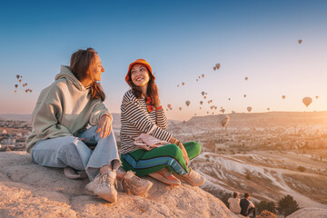 Girl friends sitting on a clifftop viewpoint and admiring view of majestic flying hot air balloons...