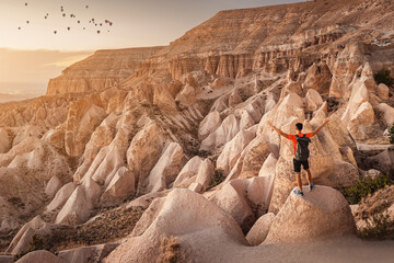 Male traveler admires geological formations of different cone shapes in Cappadocia, Turkiye and a group of flying balloons in the background at sunset