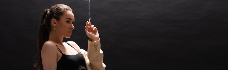 young brunette woman in beige blazer holding cigarette while looking away on black background,...