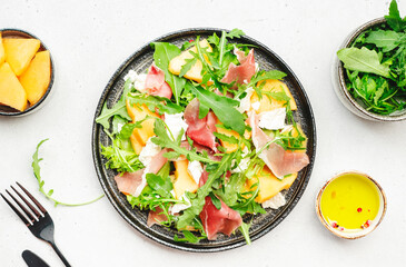 Delicious melon salad with cantaloupe, prosciutto, soft cheese and arugula on white table background, top view