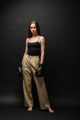 Fototapeta na wymiar full length of strong woman in top and beige pants posing in boxing gloves on black background.