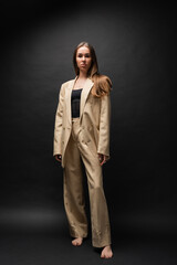 full length of barefoot woman in beige suit standing and looking at camera on black.