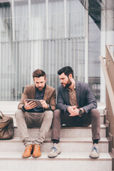 Obraz premium Two elegant contemporary bearded businessman outdoors sitting staircase sharing tablet doing online trading bitcoin cryptocurrency remote working