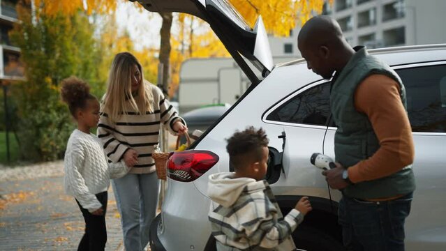 Father with his son charging their electric car, while mother with daughter packing stuff for picnic.