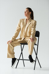 full length of brunette young woman in beige trendy suit sitting on chair on grey.