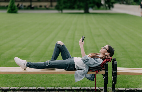 Young beautiful woman student lying on a banch in a campus listening to music or audio lectures.