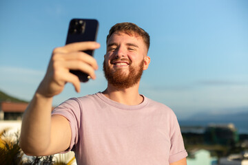 Young guy, handsome happy man is taking picture of hisself, selfie at camera of his phone, using smartphone for social media at summer in tropical exotic country. Blogger, vlog