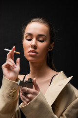 Obraz premium young brunette woman in beige blazer holding cigarette and looking at lighter on black background.