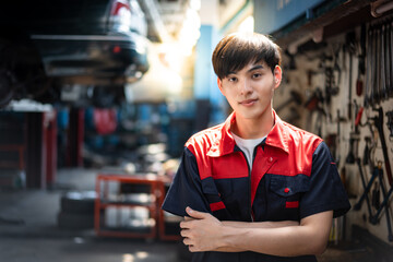 Selective focus, front view of a young handsome Asian male mechanic in red and blue uniform, standing with arms crossed and smiling at the camera with blurred lifted cars in garage in the background.