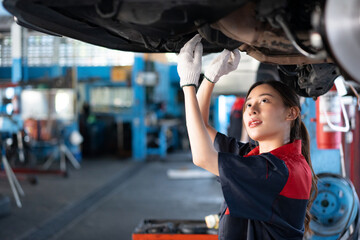 Selective focus of a young pretty Asian female mechanic in uniform, wearing work gloves examining the car bottom underneath a lifted car with a blurred garage in the background. Copy space on the left