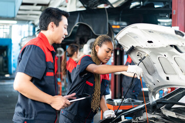 Selective focus of a young African female mechanic in uniform, checking car engine oil level with open hood, with a senior Asian male mechanic holding a tablet standing beside her in a blurred garage. - Powered by Adobe