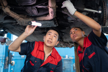 Selective focus, low angle view of mid-adult Asian male mechanic and young African female mechanic in uniform, holding a light bulb examining the car bottom underneath a lifted car in a blurred garage