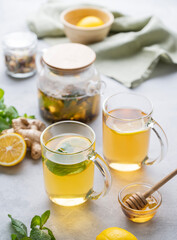 Two glasses of tea with fresh lemon, honey, mint and ginger on a light background with teapot and morning light close up.
