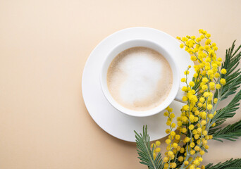 A spring bouquet with yellow mimosa flowers and a cup of coffee  cappuccino on a beige background. Concept of 8 March, happy women's day.