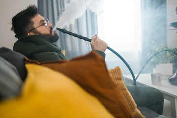 Bearded man is smoking hookah at home and blowing cloud of smoke - chill time and resting concept
