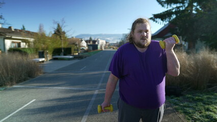 A young fat man getting back into shape building strength. Motivational concept of an overweight person listing weight dumbbells standing outside