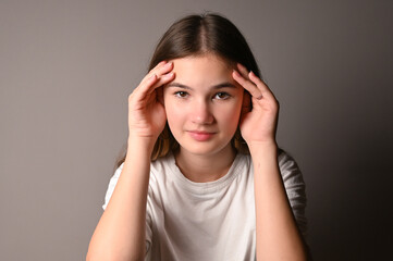 Fototapeta premium Portrait of serious teen girl on gray background. Teenage girl holds her hands to her forehead and looks at the camera