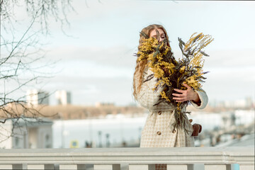 A young woman stands with a bouquet of yellow mimosa and sniffs the flowers. The concept of the...
