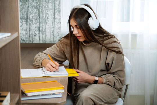Teenage girl sits at desk at home and write in a notebook. White headphones on head for listen relax music. Thinks about plans, keeps a diary book.