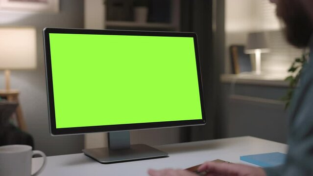Mature bearded man working on personal computer at home, cozy evening typing on keyboard, close up of green chromakey monitor