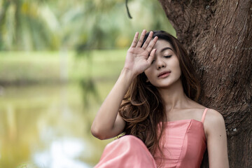 a woman in pink dress looking tired by her hand gesture and leaning to a tree