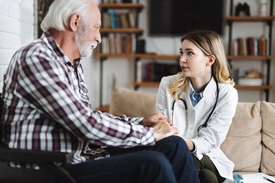 Young Nurse Talking With Sad Mature Man In A Wheelchair During Home Visit