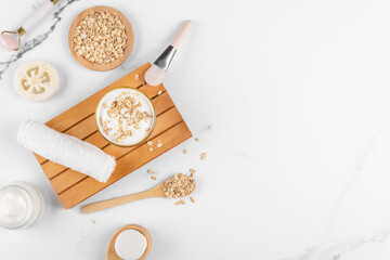Homemade facial mask or creamy scrub with cereal, made of yogurt and oats flake, home spa cosmetics on a white marble background with natural beauty treatment ingredients and towel, flat lay, top view