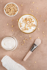 Homemade facial mask or creamy scrub with cereal, made of yogurt and oats flake, home spa cosmetics on a beige background with natural beauty treatment ingredients and brush, flat lay, top view