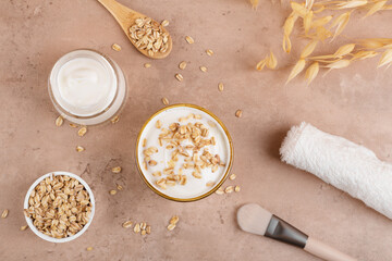 Homemade facial mask or creamy scrub with cereal, made of yogurt and oats flake, home spa cosmetics on a beige background with natural beauty treatment ingredients and towel, flat lay, top view
