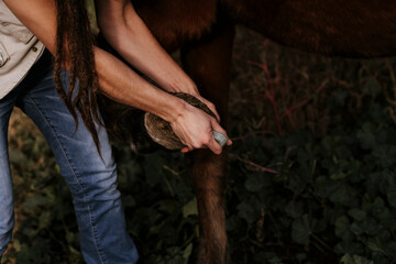 Brunette woman cleaning the hooves of a brown horse.