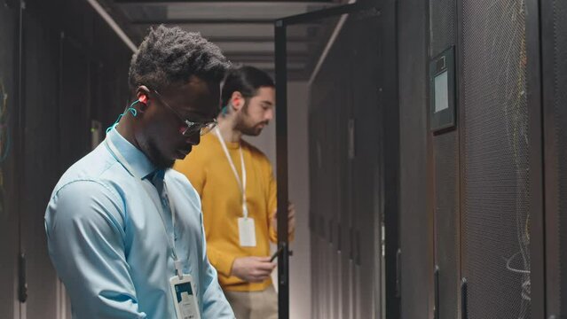 Two multiethnic male IT engineers controlling work of hardware at rack server room of data center