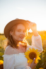 Portrait of beautiful young woman 33 years old in hat in sunflower field at sunset. Happy model in...