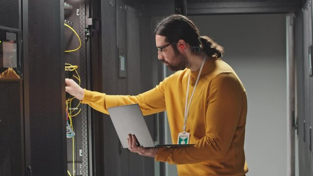 Male IT technician checking work of hardware in data center, typing program code on laptop while standing at rack server cabinet