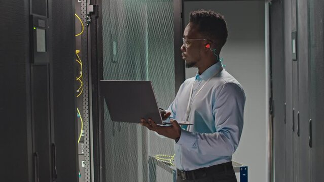 Side view medium shot of young African American male IT technician using laptop while checking work of hardware at rack server cabinet