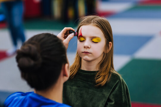 A professional make-up artist, artist paints with a brush on her face with multi-colored paints face painting, drawing, children's makeup for a little girl model. Photography, art concept, lifestyle.