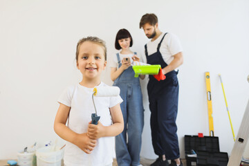 Selective focus on little cute girl holding paint roller while blurred mom and daddy standing on the background and looking for interior design idea for renovation on phone