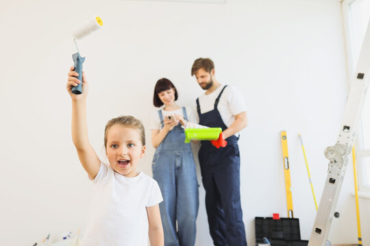 Happy overjoyed caucasian small girl raising her hand with paint roller while blurred mom and daddy standing on the background and looking for interior design idea for renovation on smart phone.